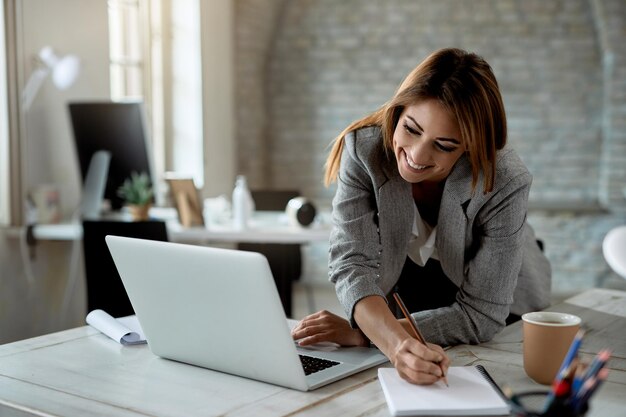 Heureuse femme d'affaires écrivant des notes tout en utilisant un ordinateur portable et en travaillant au bureau