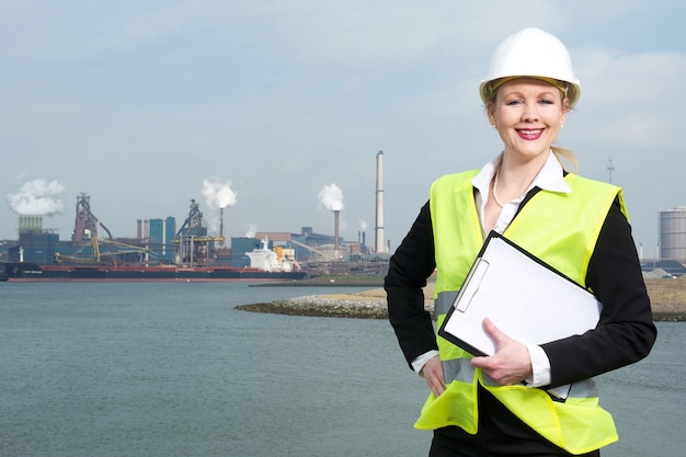 Heureuse femme d&#39;affaires en hardhat et gilet de sécurité debout à l&#39;extérieur avec le presse-papiers