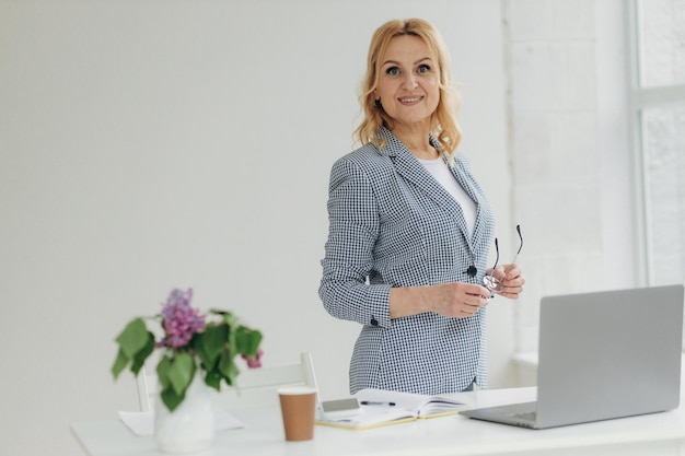 Heureuse femme d'affaires mature se penchant sur le bureau avec un ordinateur portable debout et regardant la caméra