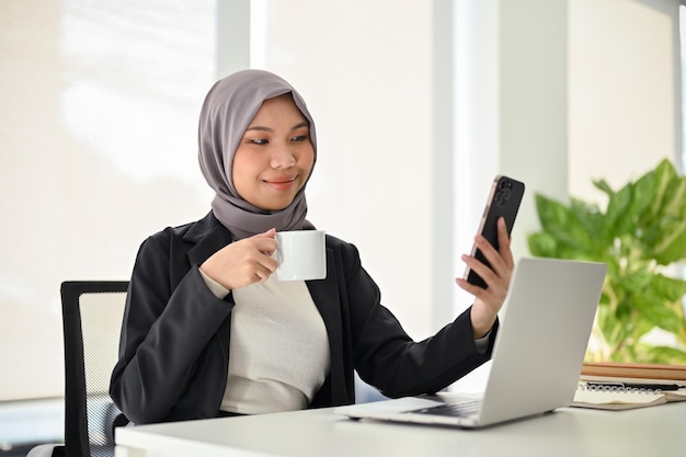 Heureuse femme d'affaires musulmane asiatique utilisant son smartphone tout en sirotant un café à son bureau