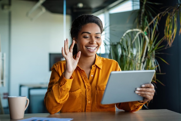 Heureuse femme d'affaires noire assise à table au bureau ayant un chat vidéo avec des partenaires commerciaux à l'aide d'une tablette numérique