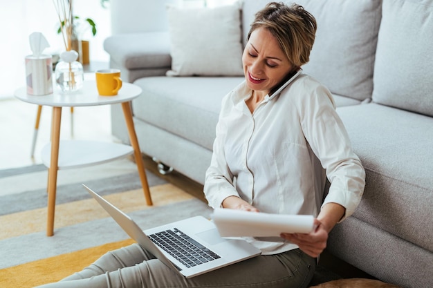 Heureuse femme d'affaires parlant au téléphone tout en utilisant un ordinateur portable et en lisant des rapports à la maison