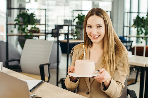 Heureuse femme d'affaires tenant un gâteau avec une bougie allumée au bureau et regardant la caméra