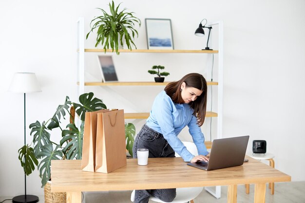 Heureuse femme d'affaires travaillant dans un bureau à domicile debout regardant son ordinateur portable entouré de sacs-cadeaux et d'emballages pour les produits en vente sur son nouveau site web