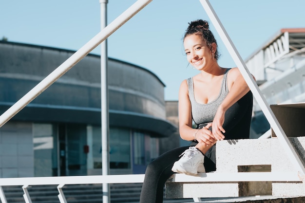 Heureuse femme africaine avec une coiffure frisée assise sur un sol urbain. Vêtements et haut de leggings. Remise en forme, souriant à la caméra. Repos après la remise en forme de l'entraînement. Tenue de sport. Mode de vie actif, pouvoir de motivation.