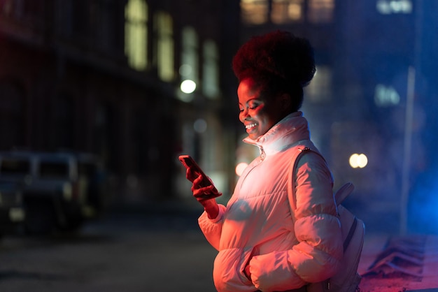 Heureuse femme africaine regardant l'écran du téléphone portable tout en se tenant à l'extérieur par une froide nuit d'hiver