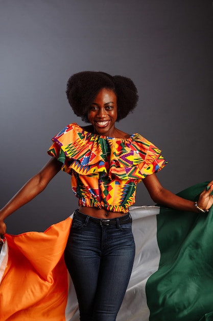 Heureuse femme africaine en vêtements nationaux souriant et posant avec un drapeau Côte d'Ivoire C te d'Ivoire isolé sur fond gris