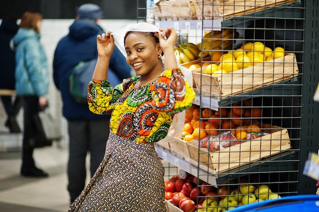 Photo heureuse femme africaine en vêtements traditionnels et voile à la recherche de produit à l'épicerie, faire du shopping dans un supermarché. les femmes afro-noires qui achètent des fruits au marché.