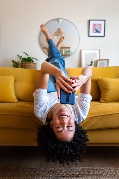 Heureuse femme afro-américaine allongée à l'envers sur le canapé envoyant des SMS à l'aide d'un téléphone portable Vérification des médias sociaux Vertical