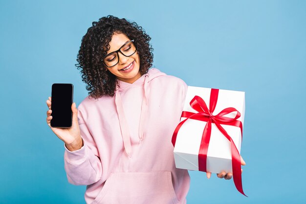 Heureuse femme afro-américaine bouclée en riant occasionnel tout en tenant présent isolé sur fond bleu. Utilisation du téléphone.