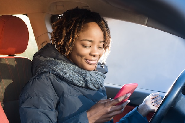 Heureuse femme afro-américaine dans un manteau vérifiant son téléphone à l'intérieur de sa voiture