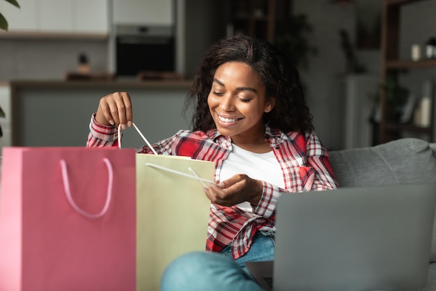 Heureuse femme afro-américaine du millénaire avec ordinateur portable assise sur un canapé et regardant dans un sac à provisions dans le salon