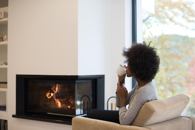 Heureuse femme afro-américaine joyeuse buvant une tasse de café relaxante au coin du feu. Jeune fille noire avec chauffage de boisson chaude se réchauffant. automne à la maison.