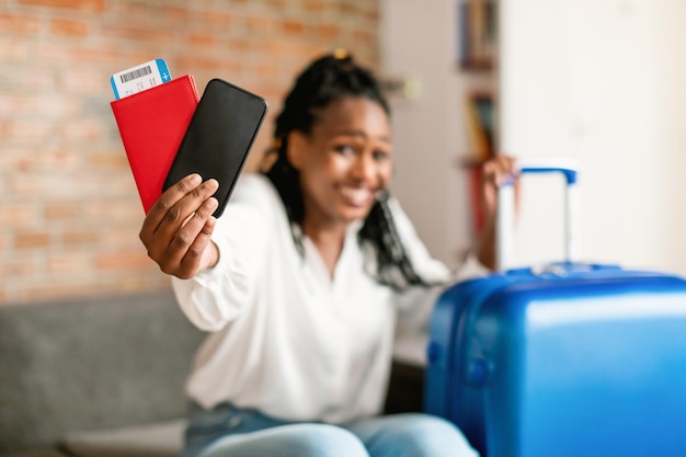 Photo heureuse femme afro-américaine montrant un smartphone avec écran vierge et tenant un passeport assis près de