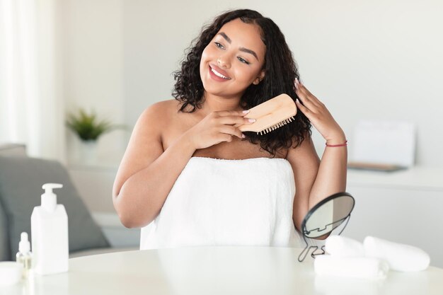 Heureuse femme afro-américaine de taille plus se brossant les cheveux après la douche assise enveloppée dans une serviette tenant une brosse en bois