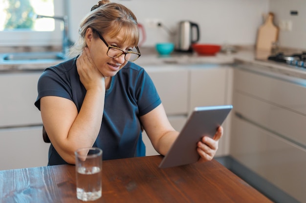 Heureuse femme d'âge moyen de 50 ans utilisant une tablette numérique assise dans la cuisine à la maison