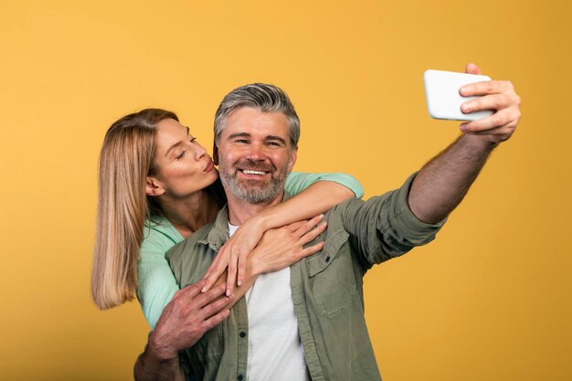 Heureuse femme d'âge moyen embrassant son mari sur la joue époux faisant selfie sur fond jaune studio shot