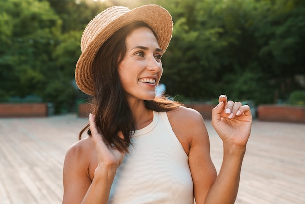 heureuse femme d'âge moyen portant un chapeau de paille souriant et regardant de côté le fond en marchant dans le parc d'été