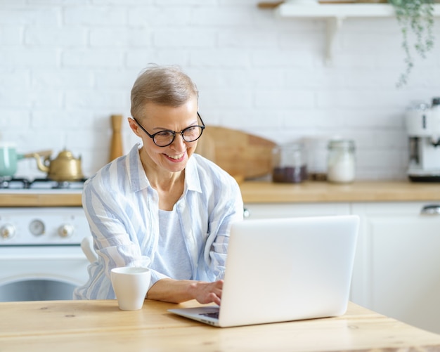 Heureuse femme d'âge moyen en train de taper quelque chose sur le clavier tout en travaillant ou en étudiant sur un ordinateur portable à la maison