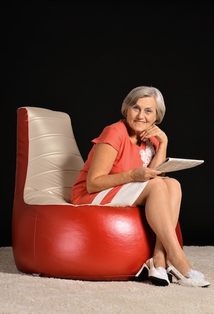 Heureuse femme âgée assise avec un journal sur un fauteuil rouge