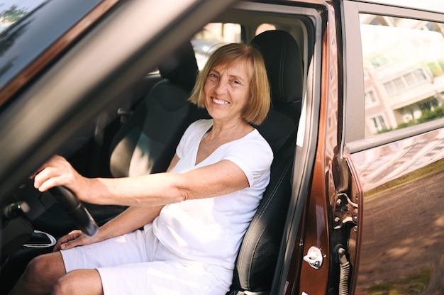 Heureuse femme âgée au volant assise dans une nouvelle voiture brune souriante regardant la caméra en profitant