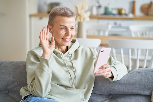 Heureuse femme âgée joyeuse ayant un appel vidéo avec un ami ou une famille sur un smartphone tout en étant assise dessus