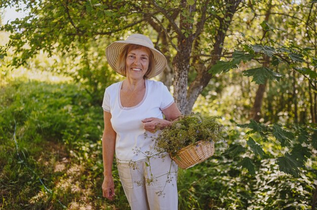 Heureuse femme âgée souriante au chapeau de paille s'amusant à poser dans le jardin d'été avec des fleurs en