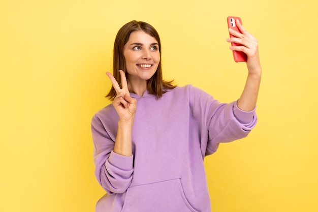 Heureuse femme agitant la main vers un smartphone et prenant un selfie montrant le signe v tout en faisant un appel vidéo