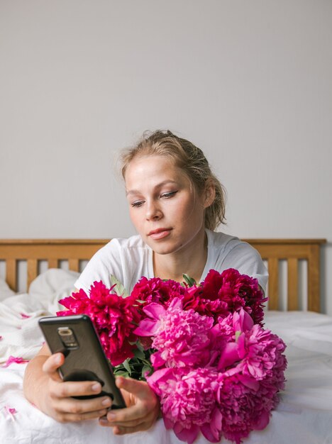 Heureuse femme allongée sur le lit en pyjama, profitant d'un bouquet de fleurs de tulipes de pivoine et utilisant un téléphone
