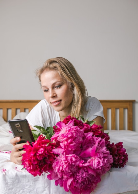 Heureuse femme allongée sur le lit en pyjama, profitant d'un bouquet de fleurs de tulipes de pivoine et utilisant un téléphone