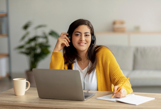 Heureuse femme arabe dans les écouteurs assis au bureau à l'aide d'un ordinateur portable et écrivant dans un cahier