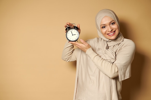 Heureuse femme arabe avec tête couverte en hijab tenant un réveil dans les mains et souriant mignon avec un sourire à pleines dents à l'avant en se tenant debout contre une surface beige avec espace copie