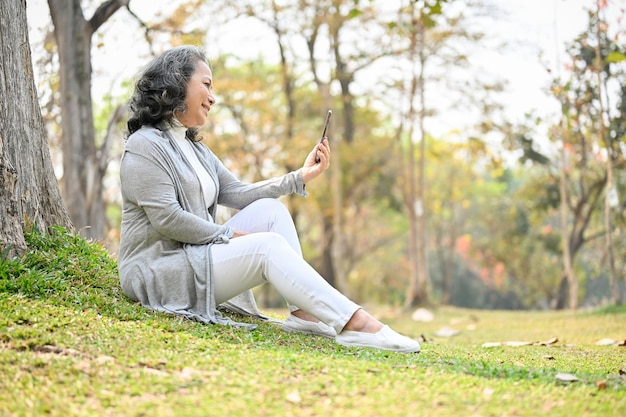 Heureuse femme asiatique de 60 ans parle lors d'un appel vidéo tout en se relaxant dans le parc