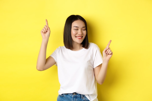 Heureuse femme asiatique dansant et s'amusant, posant en t-shirt blanc sur fond jaune.