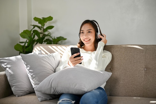 Heureuse femme asiatique écoutant de la musique à travers ses écouteurs tout en se relaxant dans le salon