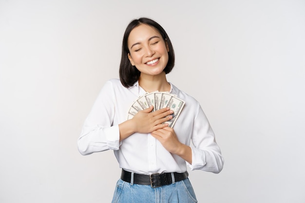 Heureuse femme asiatique étreignant des dollars d'argent et souriant satisfait debout sur fond blanc