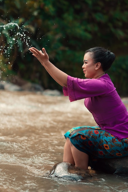 Heureuse femme asiatique jouant avec l'eau près de la rivière, assise sur le rocher et vêtue d'une robe violette