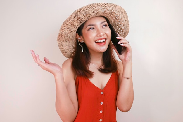 Heureuse femme asiatique portant un chapeau de paille parlant sur un téléphone portable et riant exprimant des émotions positives ayant une conversation agréable