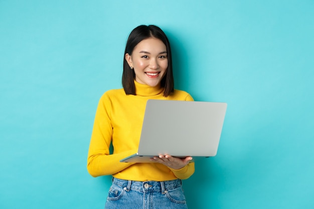 Heureuse femme asiatique en pull jaune à l'aide d'un ordinateur portable, faire des achats en ligne ou travailler, debout sur fond bleu.