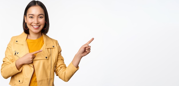 Heureuse femme asiatique souriante pointant les doigts vers la droite invitant à vérifier la vente montrant une bannière publicitaire ou un logo debout sur fond blanc