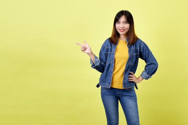 Heureuse femme asiatique en veste en jean côté pointant pour copier l'espace et en regardant la caméra sur l'espace jaune