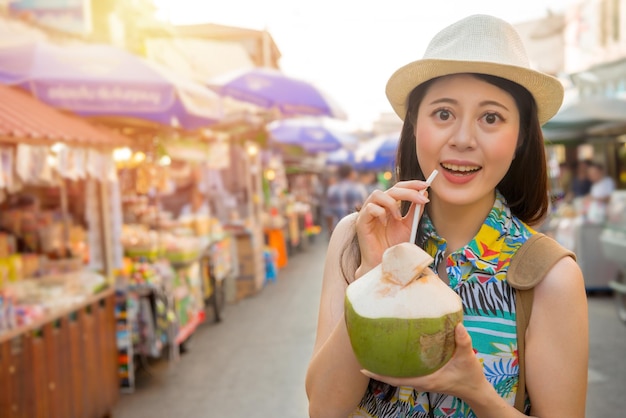 heureuse femme asiatique voyageuse tenant de l'eau de noix de coco entière debout sur la rue du marché prenant une photo souriante avec le soleil brouillant les vendeurs dans le voyage en Thaïlande vacances en Asie.