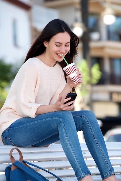 Heureuse femme assise sur un banc avec téléphone portable et boisson