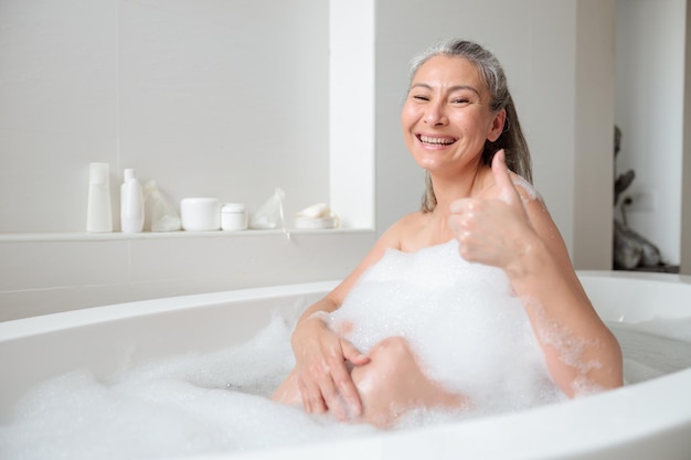 Heureuse femme assise dans la baignoire dans une salle de bain lumineuse avec de la mousse blanche, souriant et regardant la caméra