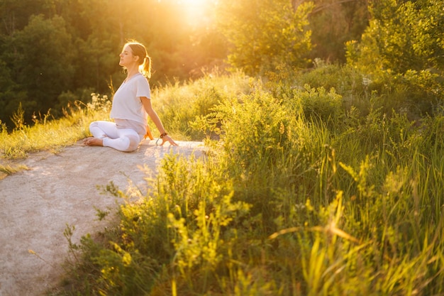 Heureuse femme assise dans la pose de lotus et faisant un virage à l'arrière à l'extérieur dans le parc sur fond de lumière du soleil