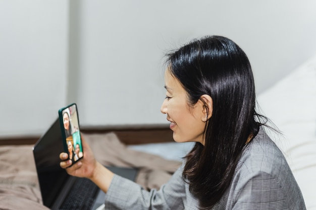 Heureuse femme assise sur le lit, faisant des appels vidéo sur smartphone.