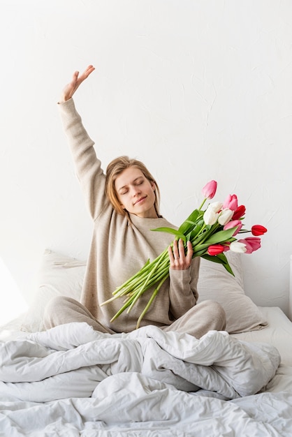 Heureuse femme assise sur le lit en pyjama, avec plaisir en appréciant les fleurs et les étirements