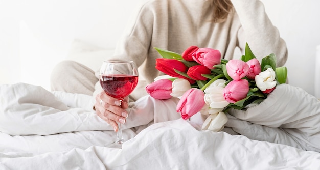 Heureuse femme assise sur le lit en pyjama, avec plaisir en appréciant des fleurs et un verre de vin rouge