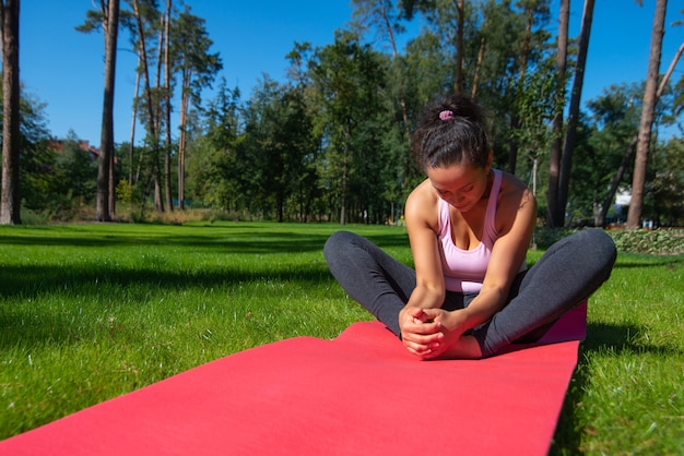 Heureuse femme assise sur un tapis de remise en forme, bénéficiant d'une séance d'entraînement dans une journée d'été ensoleillée sur fond d'une forêt de pins