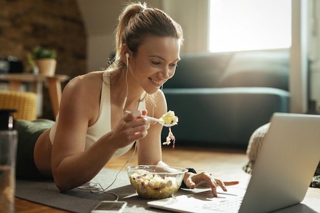 Heureuse femme athlétique profitant d'une alimentation saine et utilisant un ordinateur portable tout en se relaxant sur le sol